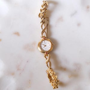 May include: A gold-tone wristwatch featuring a round white dial with thin hands and markers. The watch band is composed of interconnected infinity-shaped links and a safety chain. Two small red stones are set at the top and bottom of the watch case.