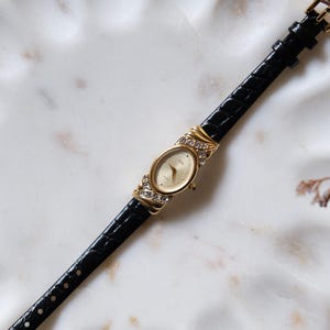 May include: A gold-tone wristwatch with an oval face and a champagne dial. The watch case is accented with clear crystals on the top and bottom lugs. It features a slim, black, textured leather strap with a gold-tone buckle closure.