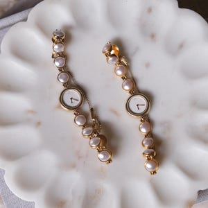 May include: A pair of gold-coloured watch earrings featuring white watch faces and pearl detailing. The earrings are displayed on a white, scalloped dish. These watch earrings are a unique jewellery item.