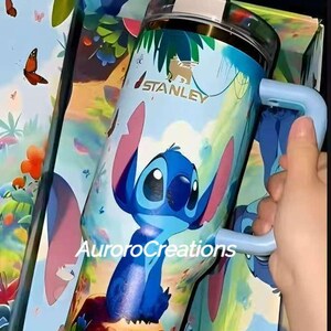 Pu&ograve; includere: Un bicchiere Stanley blu e bianco con un design di personaggi dei cartoni animati. Il bicchiere ha un manico e un coperchio con un topper per cannuccia a tema. Il testo "AuroraCreations" &egrave; visibile.