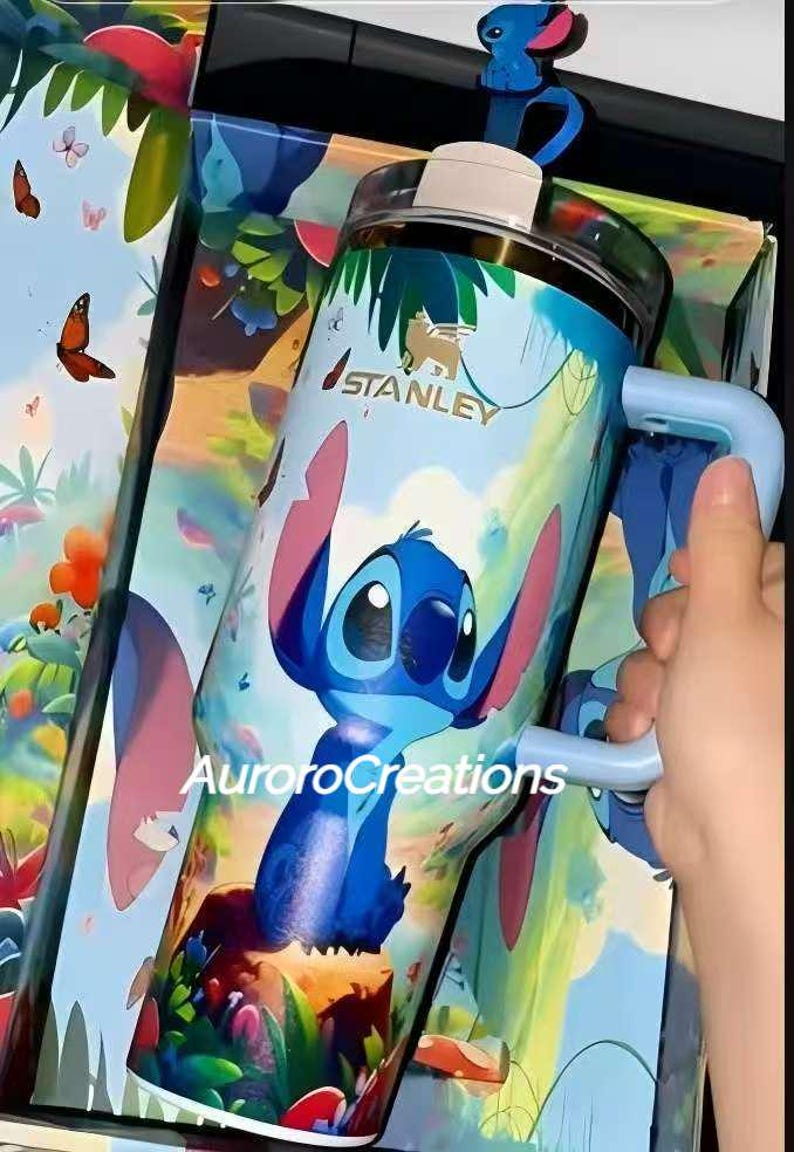 Pu&ograve; includere: Un bicchiere Stanley blu e bianco con un design di personaggi dei cartoni animati. Il bicchiere ha un manico e un coperchio con un topper per cannuccia a tema. Il testo "AuroraCreations" &egrave; visibile.