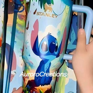 Pu&ograve; includere: Un bicchiere da viaggio Stanley con un design di personaggio dei cartoni animati blu e un manico azzurro. Il bicchiere ha un coperchio trasparente ed &egrave; decorato con fogliame tropicale e farfalle. Il testo "STANLEY" &egrave; visibile.