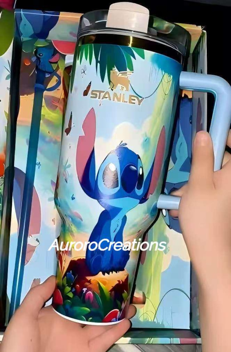 Pu&ograve; includere: Un bicchiere da viaggio Stanley con un design di personaggio dei cartoni animati blu e un manico azzurro. Il bicchiere ha un coperchio trasparente ed &egrave; decorato con fogliame tropicale e farfalle. Il testo "STANLEY" &egrave; visibile.