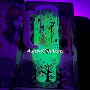 May include: A glowing green tumbler featuring characters from The Nightmare Before Christmas, including Jack Skellington and Sally. The tumbler has a white handle and the text "AuroroCreations". The design includes spiderwebs and a spooky tree.