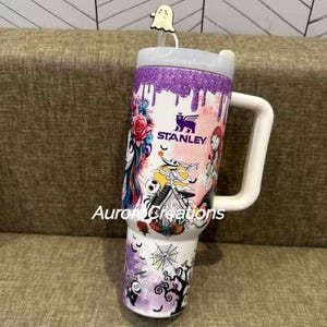 May include: A white Stanley tumbler with a large handle, featuring a colorful design with characters and elements from "The Nightmare Before Christmas." The design includes a purple glitter top, a ghost straw topper, and the text "Aurora Creations."