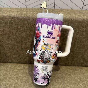 May include: A white Stanley tumbler with a large white handle, decorated with a colorful design featuring characters and elements from The Nightmare Before Christmas. The top has a purple glitter band and a ghost straw topper. The tumbler has the Stanley logo.