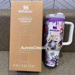 May include: A Stanley Flowstate tumbler with a white handle and lid, featuring a colorful design with characters and a purple glitter top. The box next to it has the Stanley logo and product details.