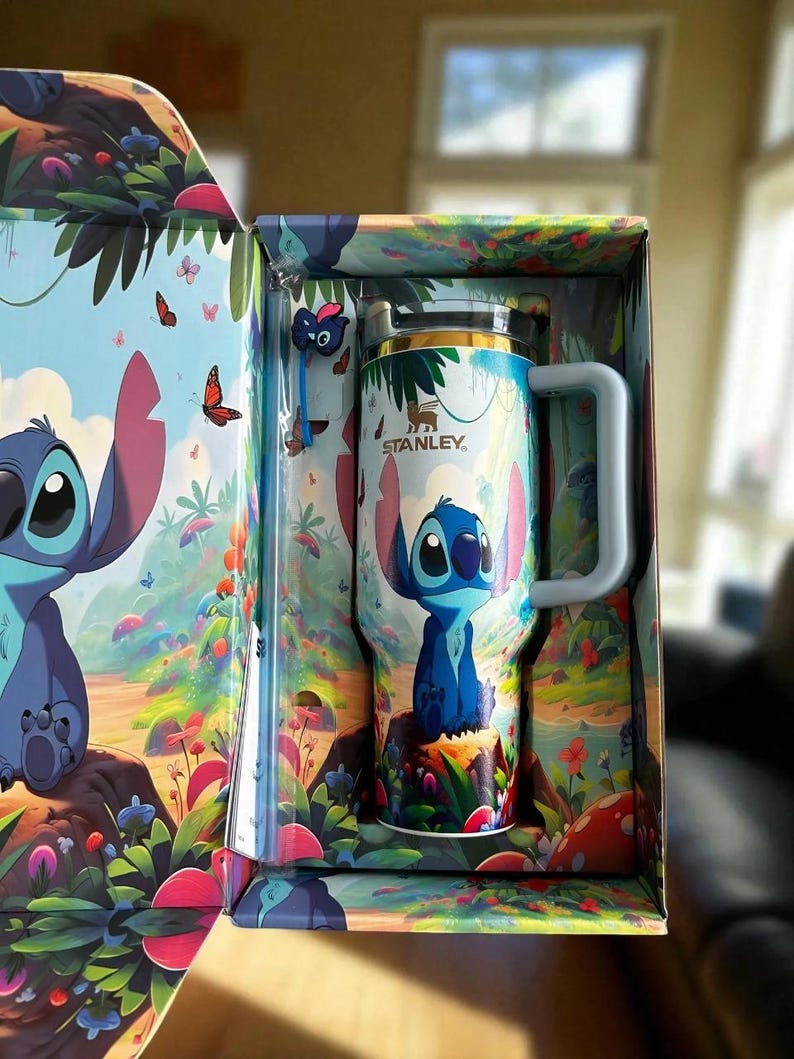 Pu&ograve; includere: Un bicchiere da viaggio Stanley con un personaggio dei cartoni animati blu, presentato in una scatola abbinata. Il bicchiere ha un manico bianco e un coperchio argentato. La scatola e il bicchiere sono decorati con un'illustrazione colorata del personaggio in un ambiente forestale stravagante con farfalle.