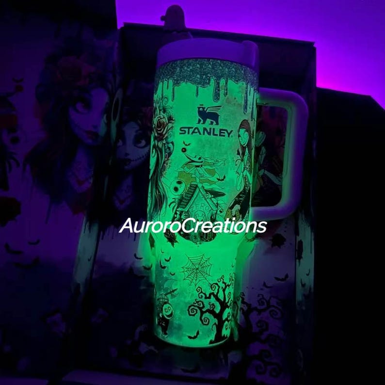 May include: A Stanley tumbler with a spooky, glow-in-the-dark design featuring characters and imagery from a Halloween-themed movie. The tumbler has a white handle and lid, with the text "Stanley" visible. The design includes a spiderweb and a tree.