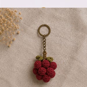 May include: A handmade keychain featuring a cluster of red crocheted grapes with green leaves. The keychain has a bronze-colored ring and chain. The grapes are made of small, round, textured stitches.