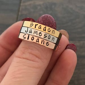 May include: Three stacked rings with the names Braden, Jameson, and Sloane engraved on them. The rings are made of different metals, with the top ring being gold, the middle ring being silver, and the bottom ring being rose gold.