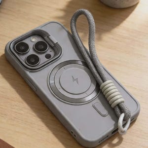 May include: A gray phone case with a magnetic charging circle and a gray lanyard. The case has a raised camera module with multiple lenses. The lanyard is attached to the case with a white loop. The phone case is on a wooden surface.