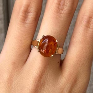 Puede incluir: Un anillo de oro con una gran piedra preciosa de color ámbar, forma ovalada y translúcida, montada en un engaste de cuatro garras. La banda es de oro pulido y se estrecha ligeramente hacia la montura central, creando un diseño clásico.