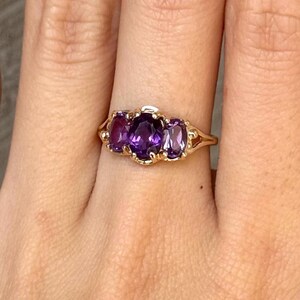Könnte beinhalten: Ein goldener Ring mit drei oval geschliffenen, violetten Amethysten, die horizontal angeordnet sind. Der mittlere Stein ist etwas größer als die beiden seitlichen Steine, alle sind in einer Krappenfassung auf einem polierten Goldring gefasst.