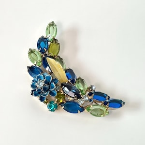 Può includere: Una spilla a forma di mezzaluna con un gruppo di strass taglio marquise nelle tonalità del blu e del verde chiaro. Il pezzo include un fiore centrale smaltato di blu con una piccola pietra chiara al centro e una singola foglia liscia giallo pallido.