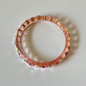 May include: A translucent pink bangle bracelet with gold-colored accents and small, iridescent rhinestones. The bracelet is circular and appears to be made of resin or a similar material. The rhinestones are evenly spaced around the bracelet.