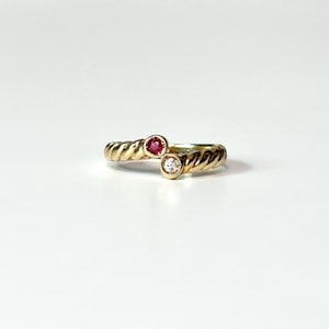 May include: A gold ring featuring a twisted rope-style band with two bezel-set stones at the center. One stone is a round, deep red ruby, and the other is a round, clear, sparkling diamond, positioned side-by-side in a bypass design.
