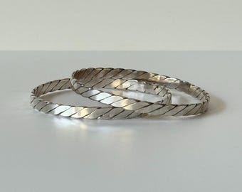 May include: Three silver-colored bangles with a braided design. The bangles are circular and appear to be made of metal. The light reflects off the surface, creating highlights and shadows. The bangles are displayed on a white surface.