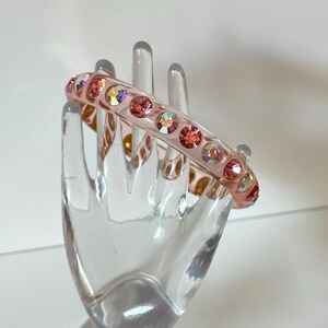 May include: A light pink hair clip adorned with a row of iridescent and pink rhinestones. The clip is curved and has a clear, glossy finish, adding a touch of sparkle and elegance. A stylish hair accessory.