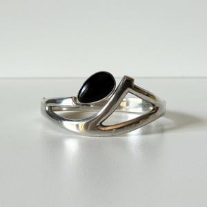 Op de afbeelding: Een zilverkleurige armband met een druppelvormige zwarte steen. De armband heeft een geometrisch ontwerp met een goudkleurig accent. De sieraden worden gepresenteerd op een wit oppervlak.