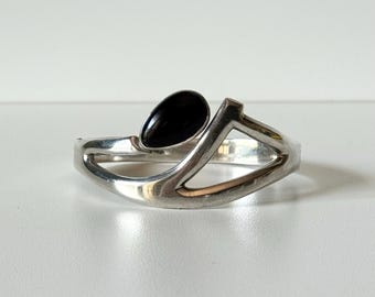 May include: A silver-toned bracelet with a teardrop-shaped black stone. The bracelet has a geometric design with a gold-colored accent. The jewelry is displayed on a white surface.