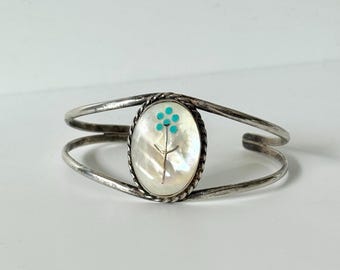 May include: A silver cuff bracelet featuring a central oval inlay of iridescent mother-of-pearl. The inlay is decorated with a small, delicate flower design composed of five tiny, bright turquoise dots and a thin silver stem.