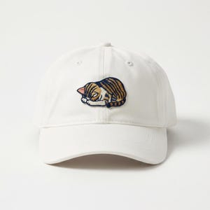 May include: White baseball cap with a patch of a sleeping cat. The cat patch features brown and tan stripes, a pink ear, and a white face. The cap is made of a soft material and has a curved brim.