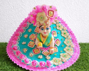 Beautiful Laddu Gopal sitting on green grass with vibrant handmade poshak, white background setup, clean and aesthetic devotional display.