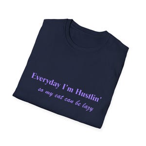 May include: Navy blue t-shirt with the text "Everyday I'm Hustlin' so my cat can be lazy" in lavender script. The shirt is folded and appears to be made of soft material. The t-shirt is a casual wear item.