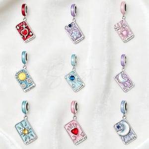 Könnte beinhalten: Eine Sammlung von neun rechteckigen Metallanhängern in Form von Tarotkarten, die jeweils ein einzigartiges Emaille-Design mit Herzen, Sternen, Sonnen und Monden in Farben wie Rot, Rosa, Blau und Violett zeigen, mit einer kleinen Öse an der Oberseite.