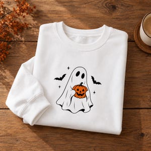Puede incluir: Una sudadera blanca de cuello redondo con un gráfico central de un fantasma blanco sosteniendo una calabaza naranja. El diseño incluye las siluetas negras de dos murciélagos volando y pequeños detalles de estrellas de cuatro puntas.
