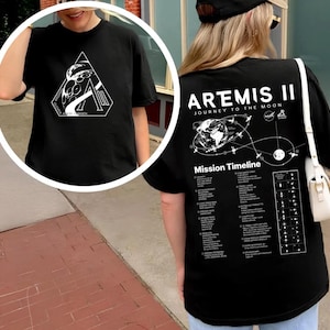 Puede incluir: Camiseta negra con gráficos blancos. La parte delantera presenta un diseño geométrico con texto. La parte trasera muestra "ARTEMIS II JOURNEY TO THE MOON" y una línea de tiempo de la misión. También es visible un logotipo de la NASA.