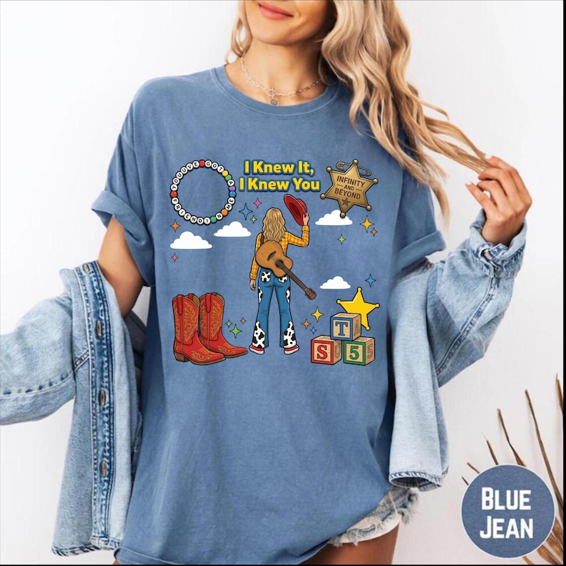 May include: A blue t-shirt featuring a graphic print of a person with blonde hair holding a red hat and a guitar, wearing cow-print trousers. The design includes red cowboy boots, a sheriff badge with text "INFINITY AND BEYOND", alphabet blocks, and the text "I KNEW IT, I KNEW YOU" with a circular beaded border.