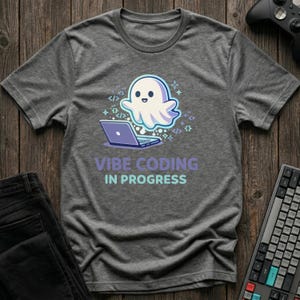 May include: A gray t-shirt with a graphic of a friendly ghost using a laptop. The text "Vibe Coding In Progress" is below the ghost and laptop. A keyboard and a game controller are visible.
