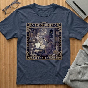 May include: Navy blue t-shirt featuring a detailed illustration of a robed figure at a desk, working on a laptop. The design includes the text "THE DEBUGGER" and "VIII ARCANA" in a decorative frame, with an owl and bookshelves.