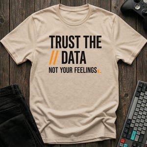 May include: A beige t-shirt with the text "TRUST THE DATA // DATA NOT YOUR FEELINGS" in black and orange. The shirt is on a wooden surface, with a pair of black jeans, a keyboard, and a game controller visible.