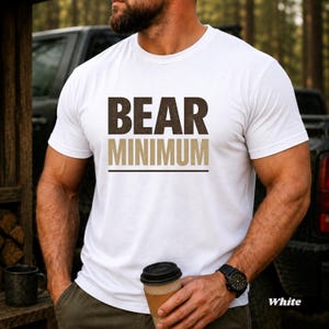 May include: White t-shirt with the text "BEAR MINIMUM" in brown and tan block letters. The shirt has a crew neck and short sleeves. A black watch is visible on the wrist. A coffee cup is held in the hand.
