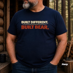 May include: Navy blue t-shirt with the text "BUILT DIFFERENT. BUILT BEAR." in white and orange lettering. The shirt is a crew neck and is being worn by a person. The word "Navy" is in the bottom right corner.
