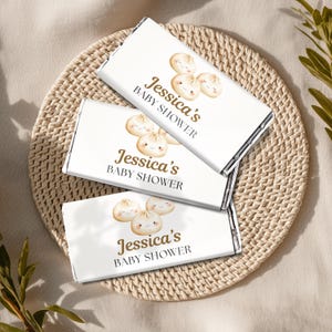 May include: Three white chocolate bar wrappers with the text "Jessica's BABY SHOWER" in brown. Each wrapper features a cartoon dumpling illustration. The bars are arranged on a woven, circular placemat.