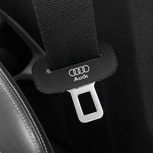 May include: Close-up of a black car seat belt with a silver buckle. The seat belt has the Audi logo and brand name printed in white on the black clasp. The seat is black leather with white stitching.