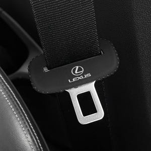May include: Close-up of a black Lexus seat belt buckle. The buckle is silver and has the Lexus logo. The seat belt is black and made of a woven material. The seat is black leather with white stitching.