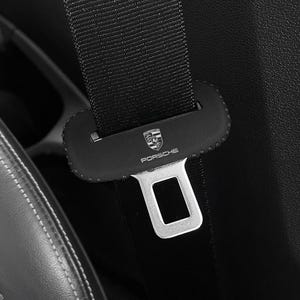 May include: Close-up of a black car seat belt with a silver buckle. The seat belt has the Porsche logo and text on the leather clasp. The seat is black with white stitching.