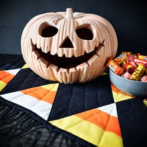 May include: A ceramic jack-o'-lantern with a wide grin sits on a black and orange candy corn patterned table runner. A bowl filled with assorted chocolate candies sits next to the jack-o'-lantern.