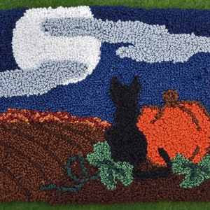 May include: A black cat and a bright orange pumpkin with green leaves sit in front of a blue sky with white clouds and a full moon. The scene is made with yarn and is a rug hooking.