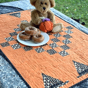 Op de afbeelding: Een oranje gewatteerde tafelloper met zwart-witte patronen. Een teddybeer met een heksenhoed en een pompoen zit op de loper, samen met een bord met donuts. Een Halloween-thema.