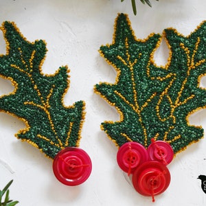 May include: Two green holly leaf ornaments with gold outlines and red button embellishments. The ornaments are made with a textured, fuzzy material.