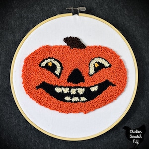 May include: A hand-stitched orange pumpkin with a black and white face and a brown stem. The pumpkin is in a white embroidery hoop.