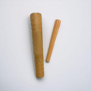 Puede incluir: Un estuche cilíndrico de madera color canela con un diseño de hoja tallada y bandas horizontales ranuradas, acompañado de una varilla de madera delgada, estriada y de color canela con un extremo cónico, ambos con textura natural.
