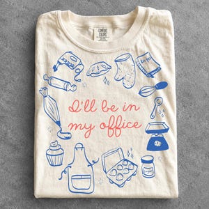 Könnte beinhalten: Ein cremefarbenes T-Shirt mit dem roten Schriftzug I'll be in my office, umgeben von blauen Strichzeichnungen von Backutensilien, darunter ein Mixer, ein Nudelholz, ein Spritzbeutel, ein Cupcake, eine Schürze, ein Eierkarton, ein Zuckerglas, eine Waage, ein Löffel, ein Schneebesen, ein Rezeptbuch, ein Ofenhandschuh und ein Kuchen.
