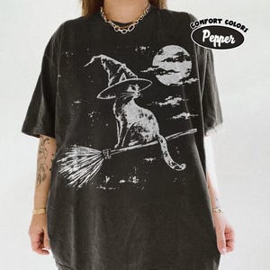 May include: A charcoal grey t-shirt featuring a white graphic print of a cat wearing a witch hat and riding a broomstick against a full moon background. The text COMFORT COLORS Pepper is printed in a black oval badge in the upper right corner.